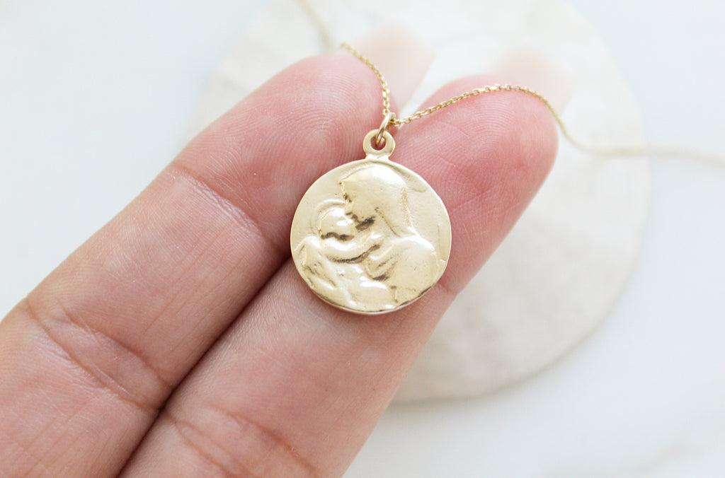 14K Gold Handmade necklace. Medallion has an image of baby Jesus being held by his mother, Mary. Mother Mary is lovingly kissing baby Jesus. Baby Jesus has his little hand on her neck. Baby Jesus has a subtle halo. The 14K Yellow Gold Necklace is on a dainty chain. This photo has the pendant placed on the hand models finger to show the size. 5/8"X9/16".