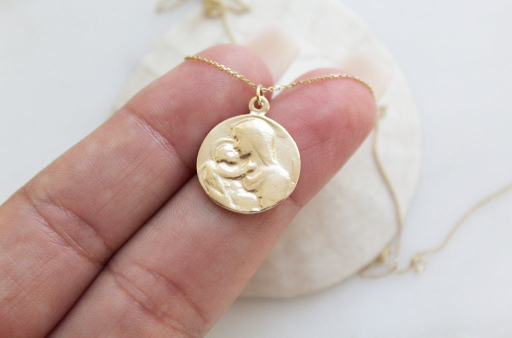 14K Gold Handmade necklace. Medallion has an image of baby Jesus being held by his mother, Mary. Mother Mary is lovingly kissing baby Jesus. Baby Jesus has his little hand on her neck. Baby Jesus has a subtle halo. The 14K Yellow Gold Necklace is on a dainty chain. This photo has the pendant placed on the hand models finger to show the size. 5/8"X9/16".